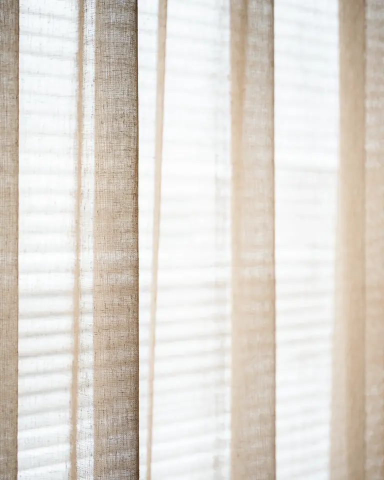 A sparse linen-texture see-through fabric against the light, with blind slats visible behind it