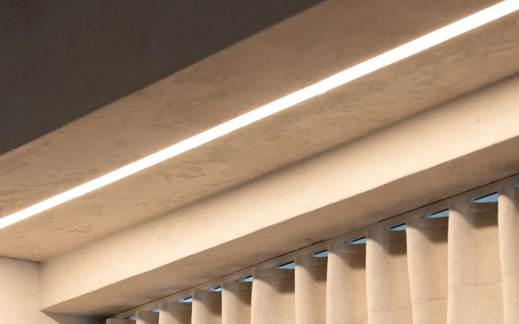 A track hidden in a plastered ceiling recess with an LED strip, carrying a linen curtain in wave pleats