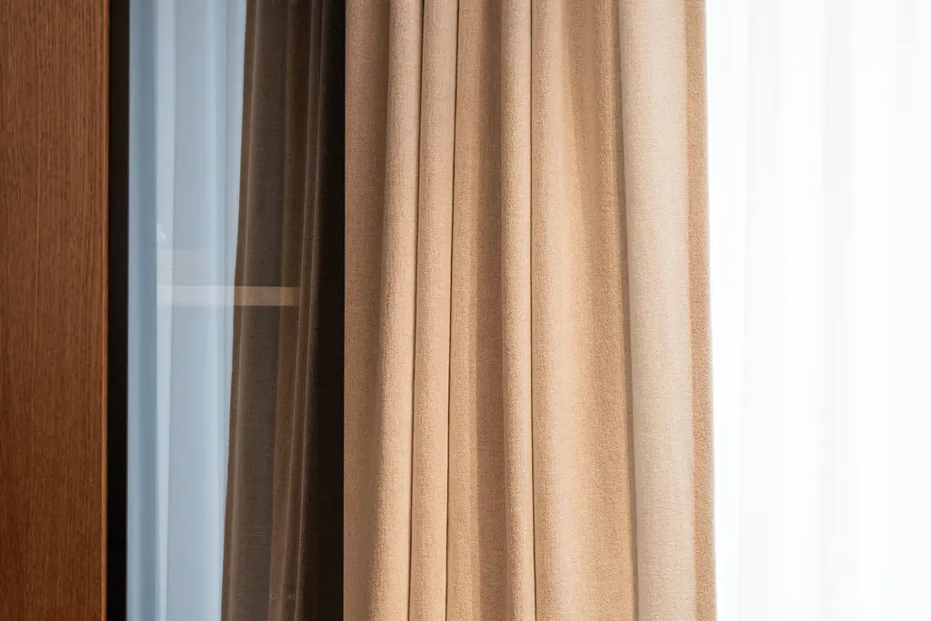 A sand-coloured linen-look curtain beside a walnut window frame, with a white sheer behind it
