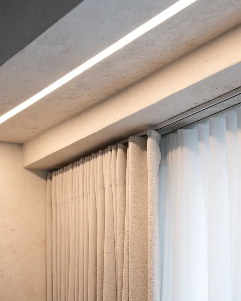 A plastered ceiling recess with a light strip, holding tracks with a linen curtain and a white sheer