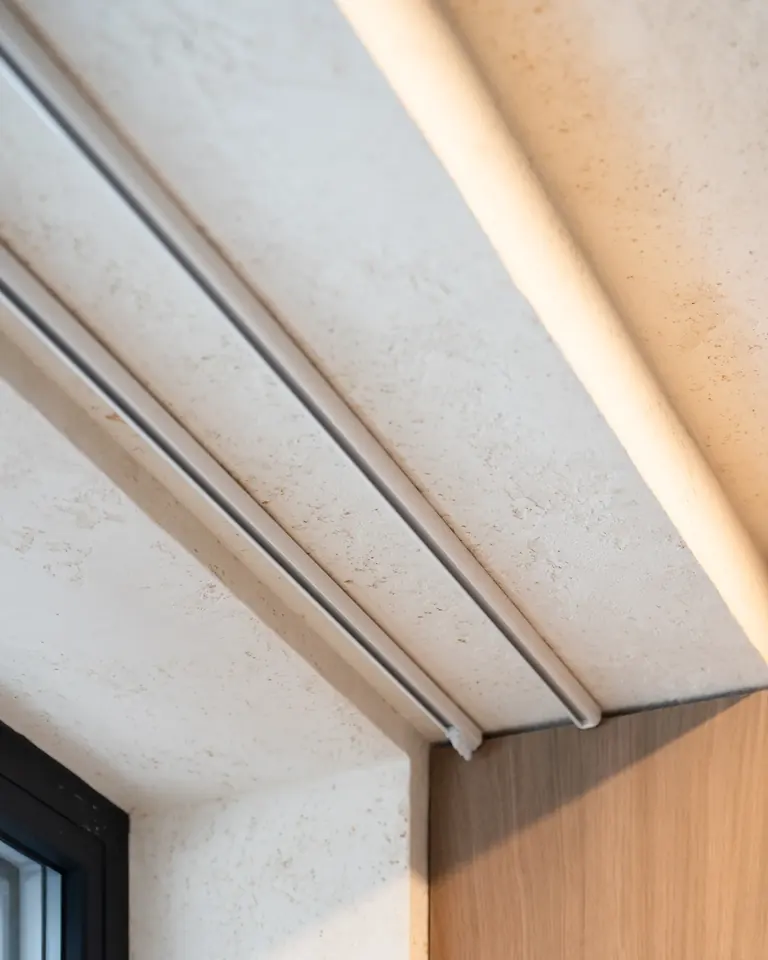 Two empty ceiling tracks in a plastered ceiling recess above a window, next to an oak finish