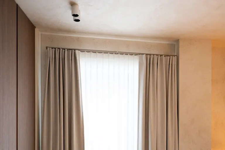 Window recess with plastered walls. Two sand-coloured curtains and a white sheer on a track below the ceiling.